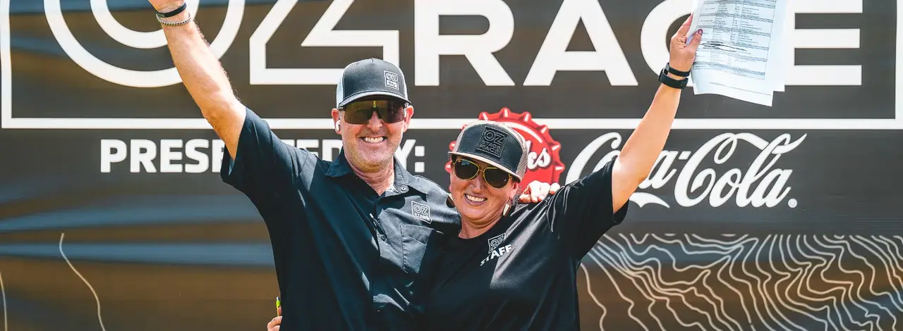 Race Director Kevin Tucker and Event Director Jamie Cathey smile and raise their arms in front of the Oz Stage Race banner.