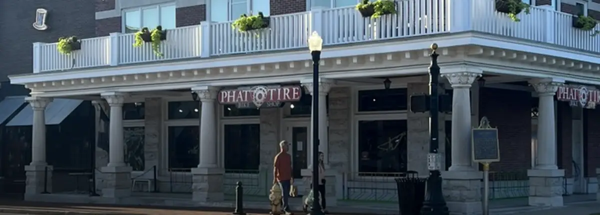 The Phat Tire Bike Shop storefront on a brick-paved corner in Bentonville, white columns and a second-story balcony under a clear sky.
