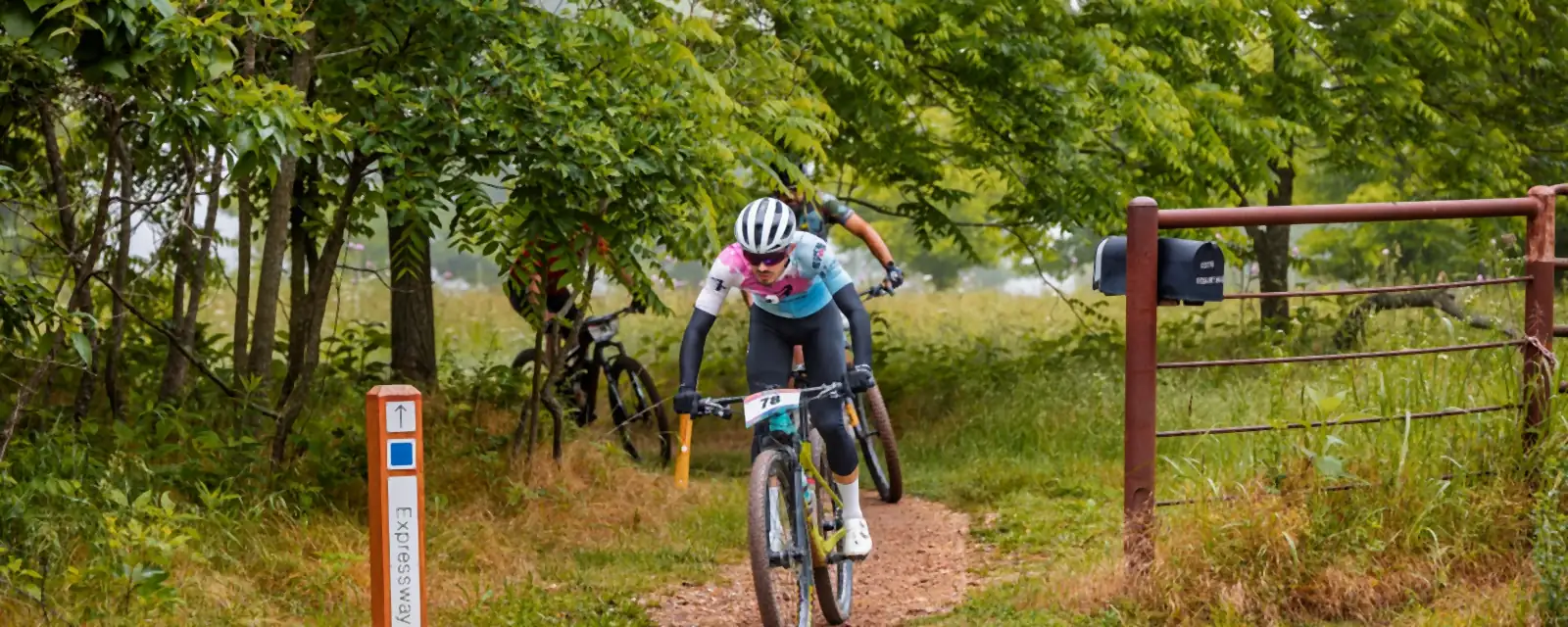 A mountain biker with race number 78 rides a dirt singletrack past an Expressway trail marker, with a rusty ranch gate and mailbox at the right and other riders behind.