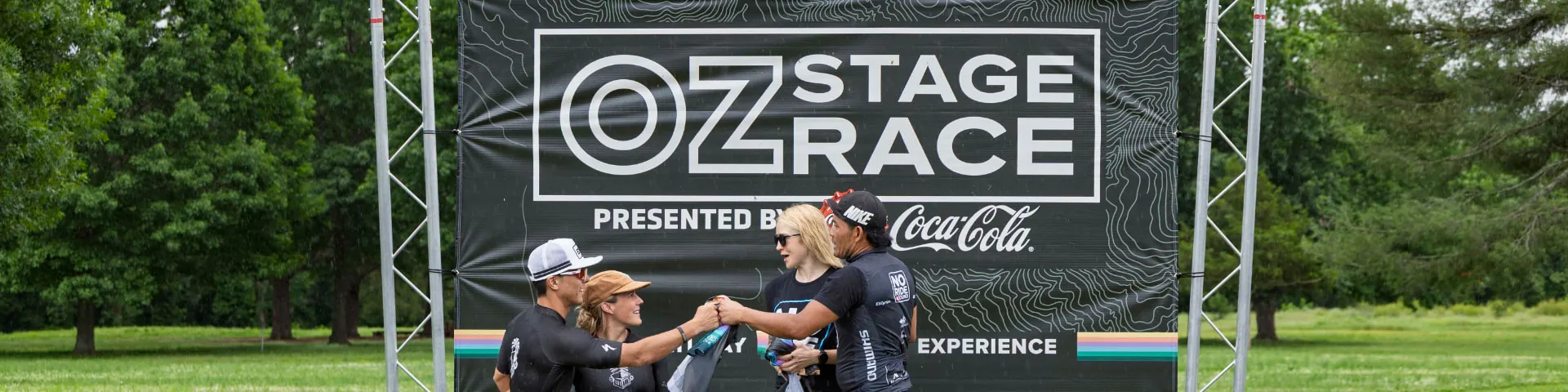 Four riders celebrate together on the podium blocks in front of the OZ Stage Race backdrop, arms around each other and drinks in hand.