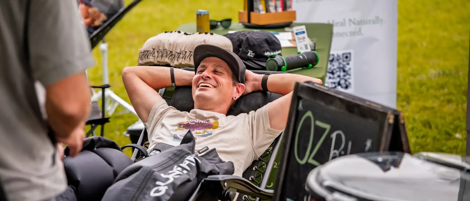 A smiling rider in a backwards cap reclines in a lawn chair with his hands behind his head and black compression boots on his legs at the race-week recovery area