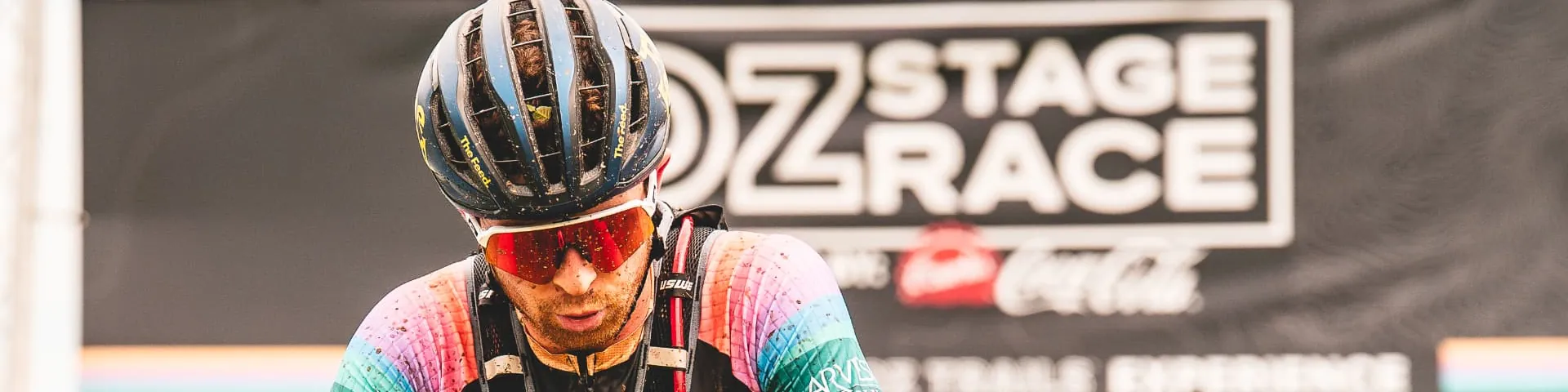 A mud-splattered racer in a rainbow-striped kit and orange-lensed glasses leans on his handlebars in front of the black OZ Stage Race banner after a stage finish.
