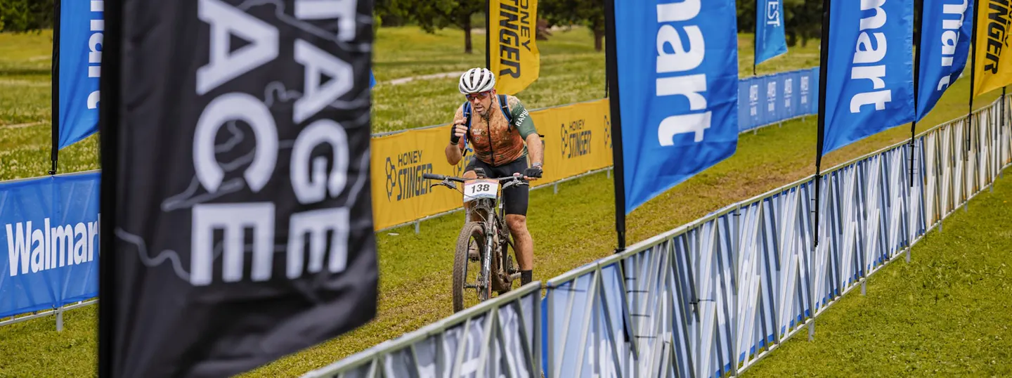 A mud-splattered rider wearing race number 138 rides toward the camera through a grassy finish chute lined with blue and yellow sponsor flags