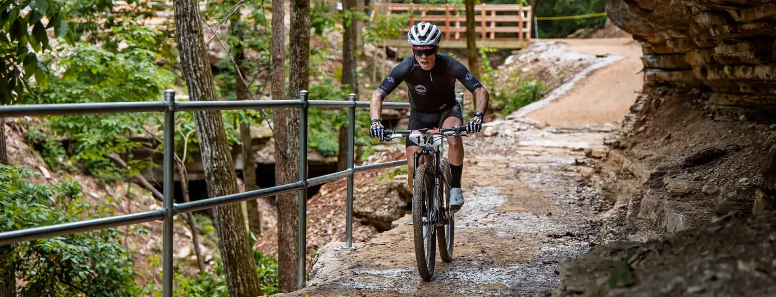 A rider in a black kit and white helmet rides toward the camera on a rocky trail cut into a cliff face, with a rock overhang at the right and a metal safety railing along a forested drop-off at the left.