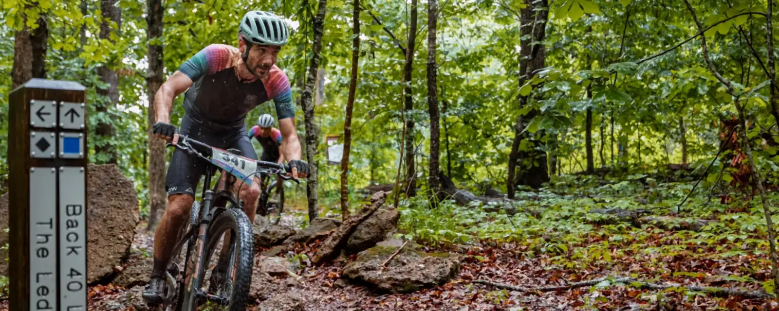 A mountain biker in helmet and racing kit rides toward the camera on a rocky forest trail, with a wooden trail sign reading Back 40 Loop and The Ledges at the left and a second rider further back.