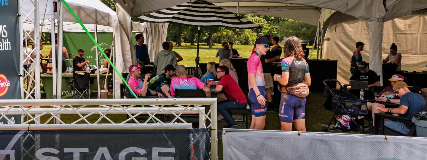 Two riders in race kits stop to talk under the event tent on the final day of the 2026 race, with the OZ Stage Race barrier running across the foreground.