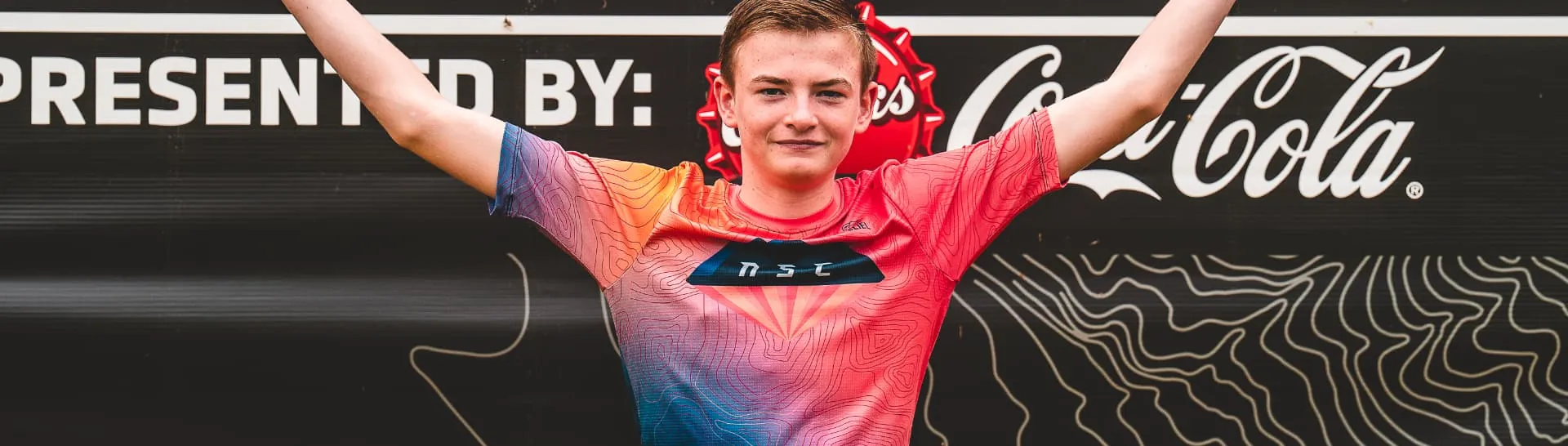 A young racer in a pink and blue jersey stands on the 2026 podium with both arms raised in front of the black OZ Stage Race backdrop.