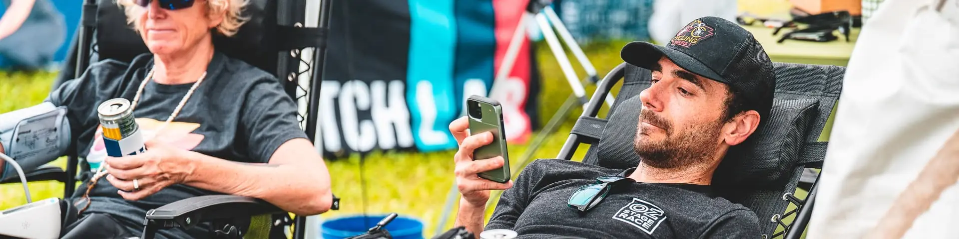 A racer in an OZ Stage Race shirt reclines in a recovery chair checking his phone while another rider holds a drink beside him at the race-week recovery area